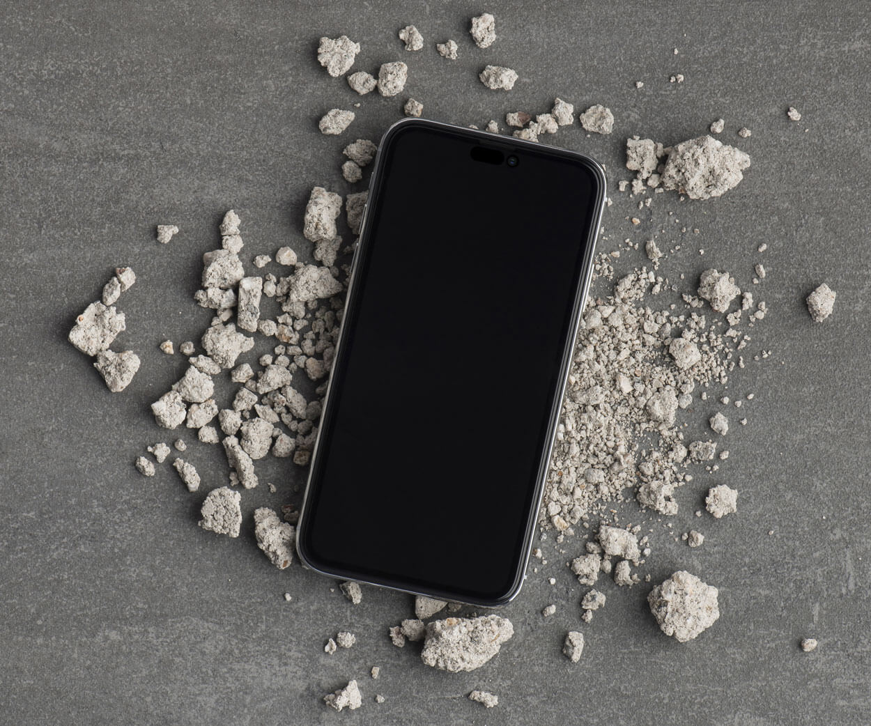 A smartphone with a black screen and dark metallic frame lies on a textured gray surface, surrounded by scattered light gray concrete or stone chips.